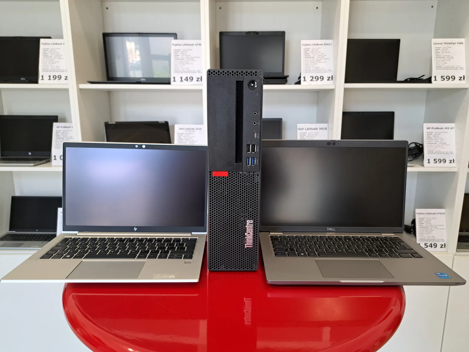 Laptopy HP EliteBook i Dell Latitude oraz komputer Lenovo ThinkCentre na ladzie warszawskiego oddziału, w tle regały z opisanym sprzętem